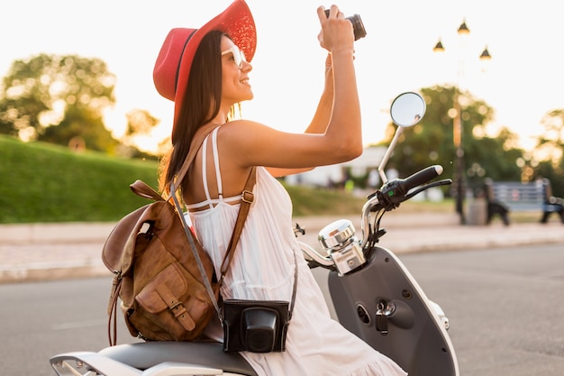 Attraente donna sorridente in sella a una moto in strada in abito stile estivo che indossa abito bianco e cappello rosso che viaggia con lo zaino in vacanza, scattare foto sulla macchina fotografica d'epoca