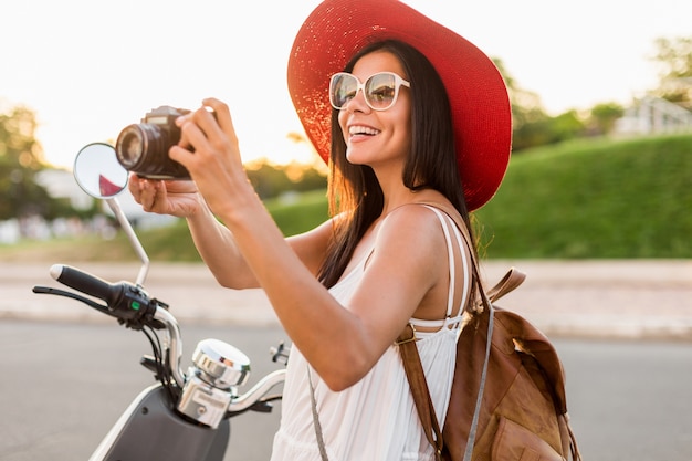 Attraente donna sorridente in sella a una moto in strada in abito stile estivo che indossa abito bianco e cappello rosso che viaggiano in vacanza, scattare foto con la macchina fotografica d'epoca