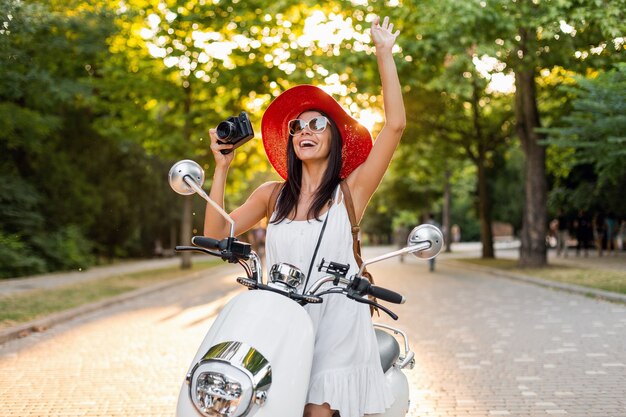 Attraente donna sorridente in sella a una moto in strada in abito stile estivo che indossa abito bianco e cappello rosso in viaggio in vacanza, scattare foto su macchina fotografica d'epoca, agitando la mano, saluto