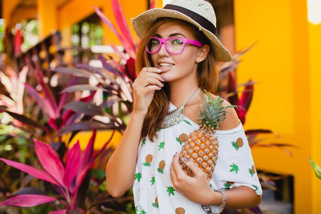 Attraente donna sorridente in vacanza in t-shirt stampata moda estate cappello di paglia, mani che tengono ananas