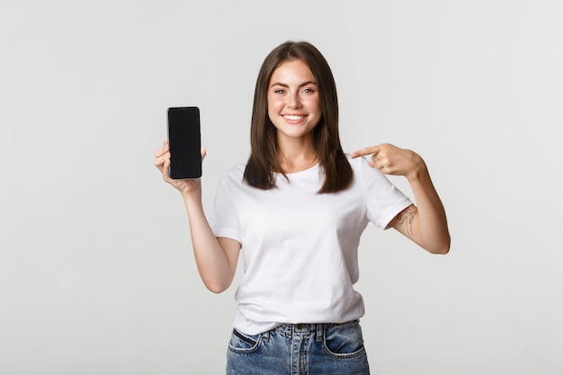 Attraente donna sorridente puntare il dito sullo schermo dello smartphone, bianco.