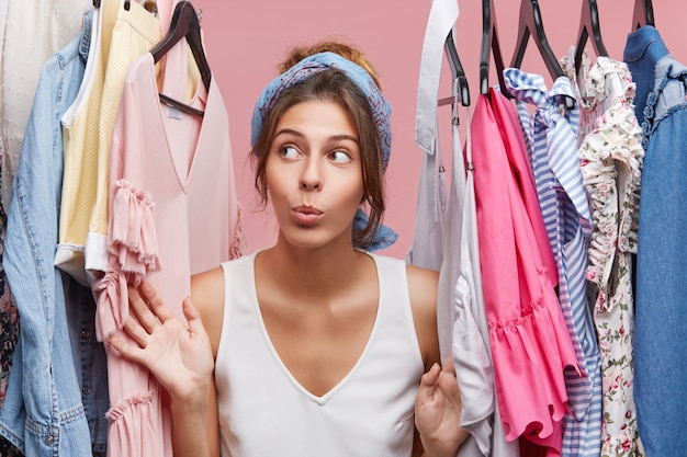 Attraente donna vestita casualmente, guardando con dubbi a parte mentre in piedi vicino a grucce con vestiti, pensando a cosa vestirsi durante un incontro di lavoro con i compagni. Donna di moda che ha molti vestiti