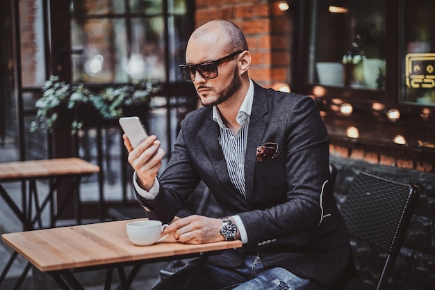 Attraente ed elegante uomo d'affari in occhiali da sole è seduto nella caffetteria fuori mentre chatta sul cellulare.