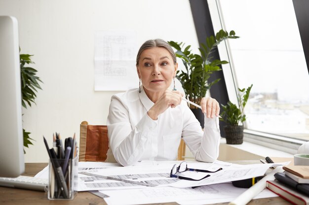 Attraente ed esperto architetto capo donna di 50 anni con i capelli grigi che studia i disegni sulla scrivania di fronte a lei, prende appunti e confronta la data con le misurazioni sul computer, avendo lo sguardo concentrato