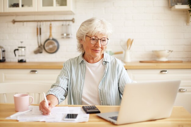 Attraente femmina matura senior positiva in bicchieri seduto al bancone della cucina davanti al computer portatile, pagare le bollette del gas e dell'elettricità utilizzando l'applicazione online, godendo della tecnologia moderna