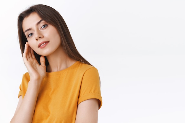 Attraente, fiduciosa donna bruna glamour in t-shirt gialla che tocca il viso perfetto senza macchia, inclina la testa leggermente aperta e la bocca rende l'espressione civettuola e sensuale, sorridente sfondo bianco