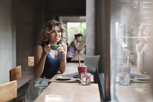 Attraente giovane biondo sognante nomade digitale seduto bere caffè caffè urbano spazio di co-lavoro guardare fuori dalla finestra sorridente sognante premuroso godere di pausa lavoro progetto freelance concetto di gig economy.