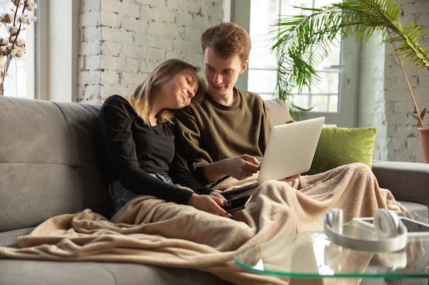 Attraente giovane coppia che utilizza dispositivi insieme, tablet, laptop, smartphone, cuffie wireless. Comunicazione, concetto di gadget. Tecnologie che collegano le persone in autoisolamento. Stile di vita a casa.