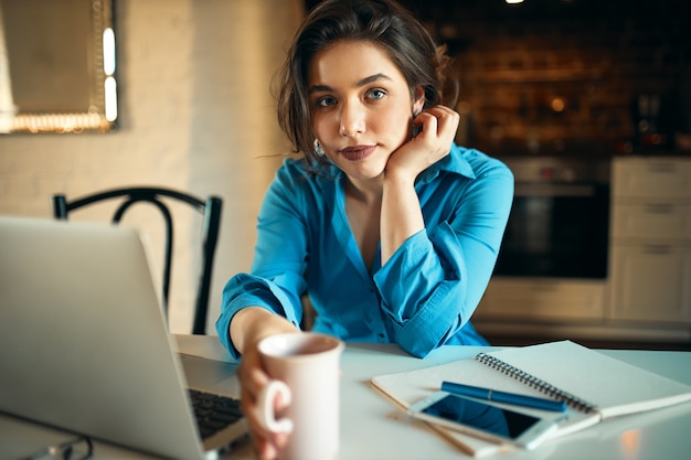 Attraente giovane dipendente di sesso femminile utilizzando laptop per lavoro remoto seduto alla scrivania con cellulare e tazza, bere caffè, fare report. Ragazza carina studentessa che studia online sul computer portatile a casa