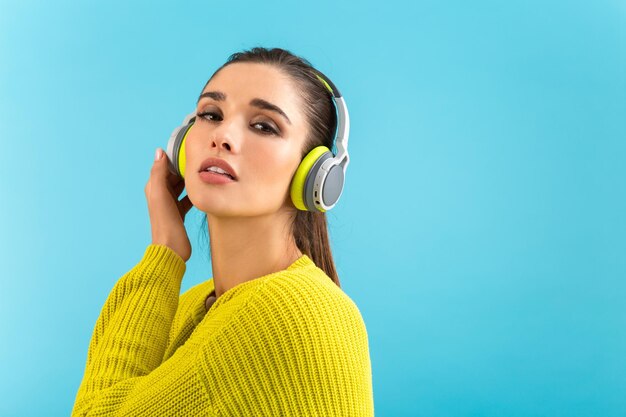 Attraente giovane donna alla moda che ascolta la musica in cuffie wireless felice indossando maglione lavorato a maglia giallo stile colorato moda in posa isolato su sfondo blu