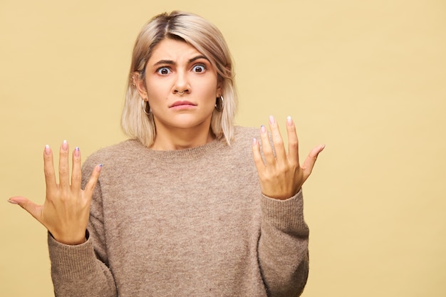 Attraente giovane donna alla moda con l'acconciatura bob bionda gesticolando emotivamente con espressione facciale indignata, scrollando le spalle, sentendosi confusa o indignata. Reazione umana e sentimenti