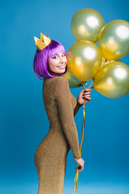 Attraente giovane donna allegra in vestito alla moda di lusso che celebra la grande festa. Palloncini dorati, corona, capelli viola tagliati, trucco luminoso, sorridente, festività natalizie.