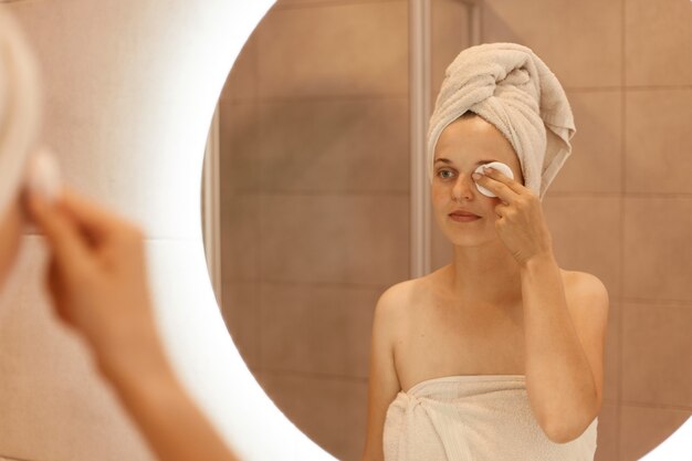 Attraente giovane donna caucasica adulta in asciugamano sui capelli guardandosi allo specchio e pulendo il viso con un batuffolo di cotone, in piedi in bagno dopo aver fatto la doccia.