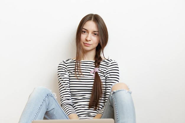 Attraente giovane donna che indossa t-shirt a maniche lunghe a righe e jeans stracciati seduti sul pavimento