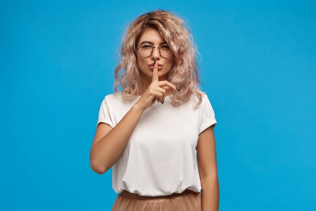 Attraente giovane donna con i capelli ricci disordinati che indossa occhiali rotondi e top oversize che chiede di tacere, tenendo l'indice sulle labbra, emettendo un suono smorzato. Segretezza