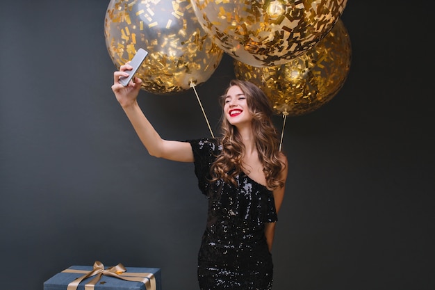 Attraente giovane donna con l'acconciatura riccia che fa selfie in camera con interni neri durante la festa. Raffinata ragazza bionda caucasica festeggia il compleanno e ride.