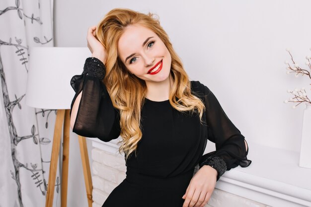 Attraente giovane donna con un bel sorriso, toccando sensualmente i suoi capelli biondi ondulati in camera bianca moderna. Indossare un abito nero alla moda, rossetto rosso, trucco leggero.