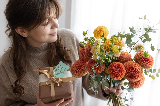 Attraente giovane donna con un regalo per la festa della mamma e un mazzo di fiori di crisantemo nelle sue mani.