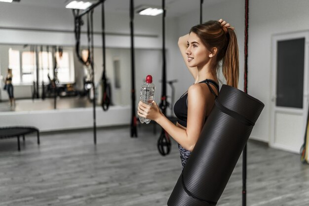 Attraente giovane donna con un tappetino da yoga e una bottiglia d'acqua in una moderna palestra leggera