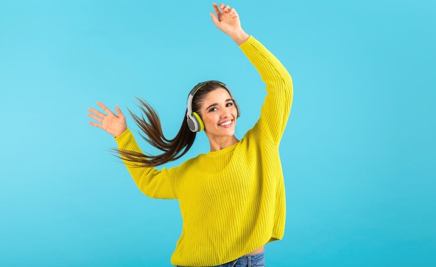 Attraente giovane donna elegante che ascolta la musica in cuffie senza fili felice indossa maglione giallo lavorato a maglia stile colorato moda in posa isolato su sfondo blu agitando la coda di capelli lunghi