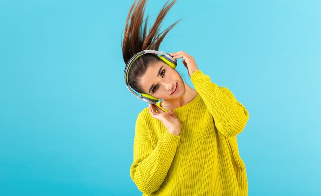 Attraente giovane donna elegante che ascolta la musica in cuffie senza fili felice indossa maglione giallo lavorato a maglia stile colorato moda in posa isolato su sfondo blu agitando la coda di capelli lunghi