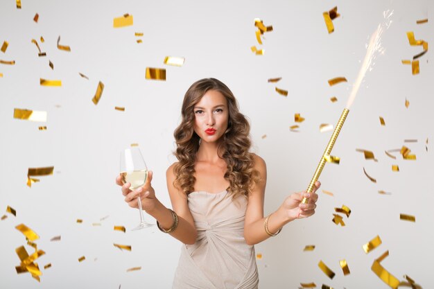 Attraente giovane donna elegante che celebra il nuovo anno, bevendo champagne, coriandoli dorati che volano, sorridendo felice, isolato, con indosso un abito da festa