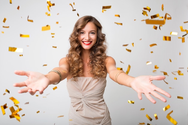 Attraente giovane donna elegante che celebra il nuovo anno, coriandoli dorati che volano, sorridendo felice, isolato, vestito da festa, trucco e acconciatura