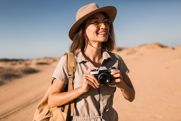 Attraente giovane donna elegante in abito color cachi nel deserto, viaggiando in Africa in safari, indossando cappello e zaino, prendendo foto con la macchina fotografica d'epoca