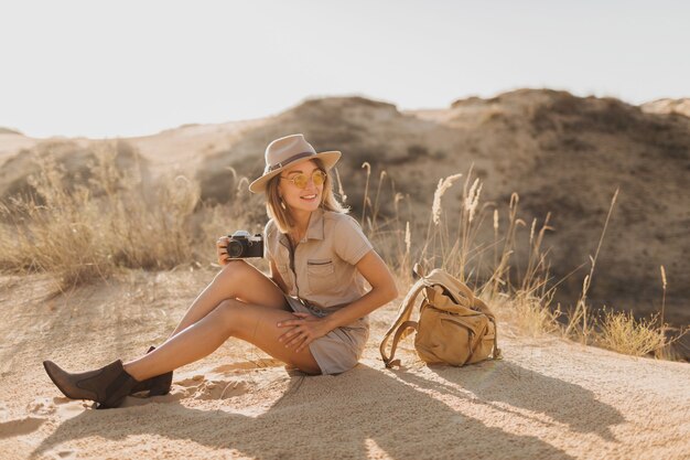 Attraente giovane donna elegante in abito color cachi nel deserto, viaggiando in Africa in safari, indossando cappello e zaino, prendendo foto con la macchina fotografica d'epoca