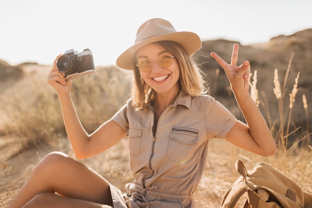 Attraente giovane donna elegante in abito color cachi nel deserto, viaggiando in Africa in safari, indossando cappello e zaino, prendendo foto con la macchina fotografica d'epoca