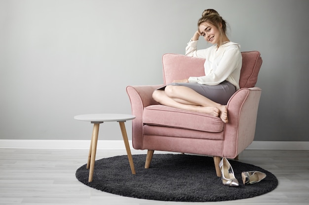 Attraente giovane donna europea alla moda con chignon e piedi nudi comodamente seduti in poltrona rosata e sorridente, godendosi il tempo libero da sola, eleganti scarpe col tacco alto sul tappeto