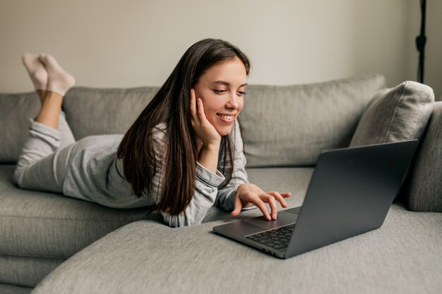 Attraente giovane donna europea con lunghi capelli scuri che indossa un pigiama che lavora a casa con il computer portatile durante la quarantena.