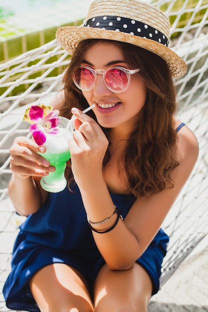 Attraente giovane donna in abito blu e cappello di paglia che indossa occhiali da sole rosa, bere cocktail alcolici in vacanza e seduto in amaca