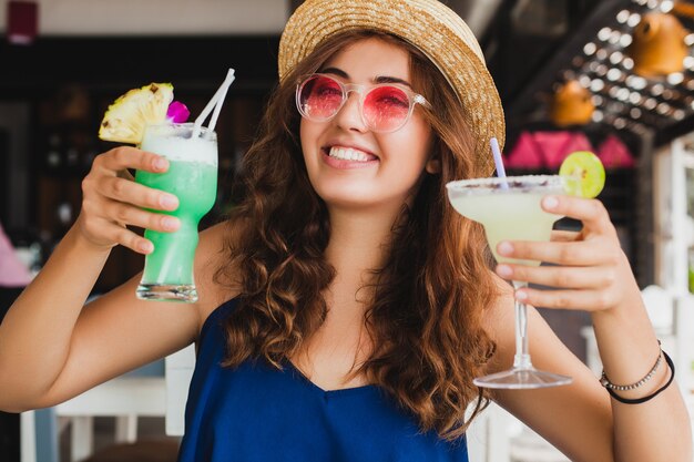 Attraente giovane donna in abito blu e cappello di paglia che indossa occhiali da sole rosa, bere cocktail alcolici in vacanza tropicale e seduto al tavolo al bar