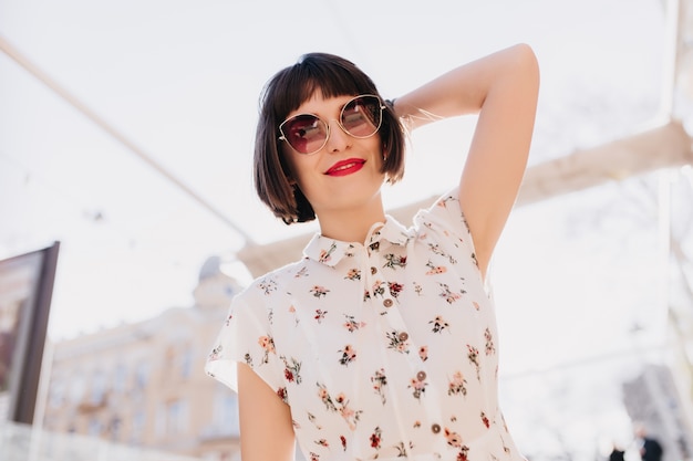 Attraente giovane donna in eleganti occhiali da sole in posa con un sorriso romantico. Ritratto all'aperto di signora europea dai capelli corti in abito bianco agghiacciante per strada.