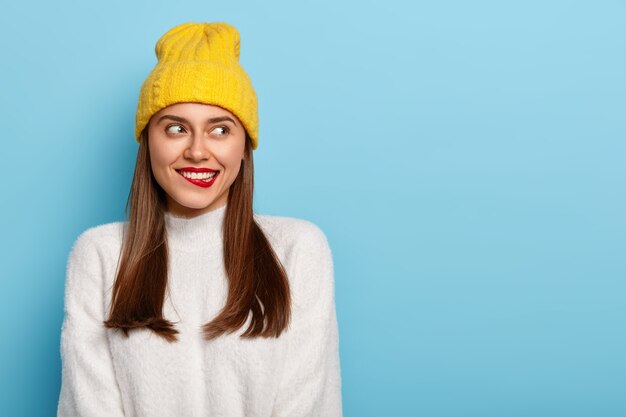 Attraente giovane donna morde le labbra rosse, pensa a qualcosa, distoglie lo sguardo, ha i capelli lisci e scuri, indossa un cappello giallo, un maglione bianco caldo