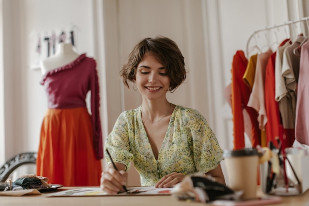 Attraente giovane donna riccia in elegante abito floreale con scollo a V sorride sinceramente, tiene la penna e posa nell'ufficio dello stilista