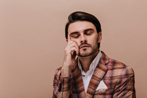 Attraente giovane gentiluomo con gli occhi chiusi, capelli castani e setole, indossa una camicia bianca e una giacca calda scura in posa contro il muro beige e tocca il viso con una mano