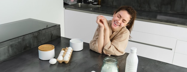 Attraente giovane ragazza allegra che cuoce in cucina preparando un impasto tenendo il ricettario con idee