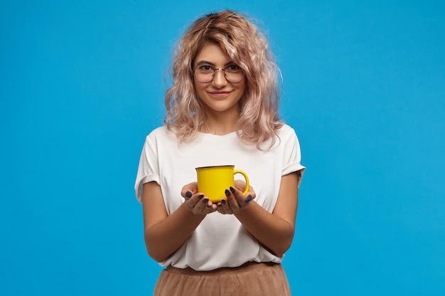 Attraente giovane segretaria femminile dall'aspetto amichevole che indossa t-shirt bianca e occhiali da vista in posa al muro blu con una tazza gialla sulle mani, offrendoti tè o caffè appena fatto