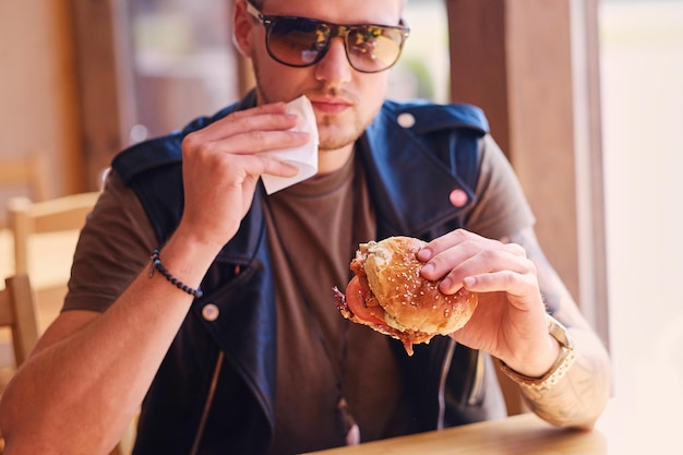 Attraente hipster vestito con una giacca di pelle che mangia un hamburger vegano.