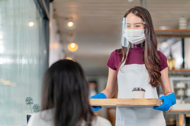 Attraente lavoratrice asiatica del caffè indossa maschera e guanti dando caffè caldo da asporto al cliente