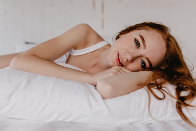Attraente modello femminile dai capelli rossi sdraiato sul cuscino e alla ricerca. Foto interna di bella donna felice con riccioli di zenzero.