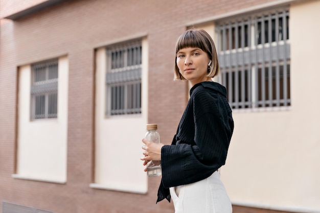 Attraente modello femminile in abiti eleganti che si gode la giornata estiva La foto all'aperto di una splendida donna dai capelli corti tiene in mano una bottiglia d'acqua e posa davanti alla telecamera con un bel sorriso