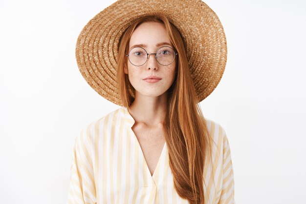 Attraente ragazza alla moda hipster con capelli rossi e lentiggini in occhiali alla moda cappello di paglia e camicetta gialla carina sorridente con espressione spensierata compiaciuta che frequentano lezione interessante nella caffetteria