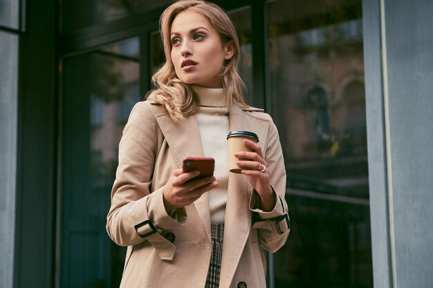 Attraente ragazza bionda casual in elegante trench con caffè da asporto e cellulare all'aperto