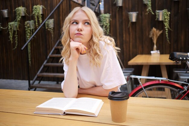 Attraente ragazza bionda casual premurosamente distogliendo lo sguardo con libro e caffè nel cortile del caffè della città