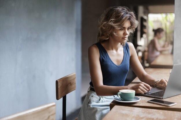 Attraente ragazza bionda freelance lavorando laptop prendendo appunti notebook sedersi da solo caffè vicino alla finestra bere caffè, scrittore professionista fare post in linea blog, preparare file riunione di lavoro dopo pranzo.