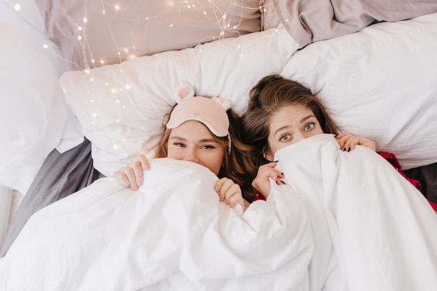 Attraente ragazza bionda in maschera da notte rosa che si nasconde sotto la coperta. Foto di interni di due raffinate sorelle che scherzano durante il servizio fotografico mattutino.