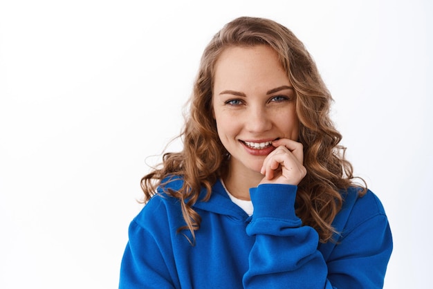 Attraente ragazza bionda strizzando gli occhi toccando le labbra e sorridendo incuriosita vedendo qualcosa di interessante guardando sfondo bianco premuroso